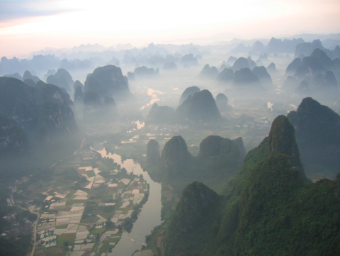 An aerial view shows a landscape of karst mountains shrouded in mist, with rice paddies and rivers winding through the valleys.