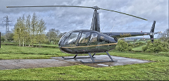 A black Robinson R44 helicopter sits on a concrete helipad surrounded by green grass and trees under a cloudy sky.