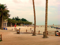 A beach scene with a wooden boardwalk in the foreground, leading to a sandy beach filled with people, palm trees, and boats on the water.