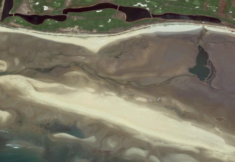 Aerial view of a coastal landscape showing a sandy beach, a tidal flat, and a golf course with ponds in the upper section of the image.