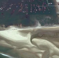 An aerial view shows a coast with several small structures on the water, and a sandy beach with patterns created by the water flowing over the sand.