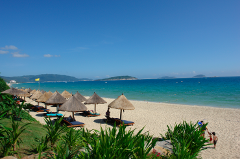 A sunny beach scene with straw umbrellas and lounge chairs, bordered by green plants, with mountains visible in the distance.