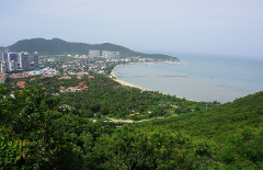 An aerial shot showcases a coastal city with lush greenery, modern buildings, a sandy beach, and a long pier extending into the ocean.