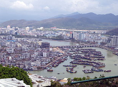 A scenic view showcases a bustling harbor filled with boats, surrounded by a densely built city and majestic mountains in the background.