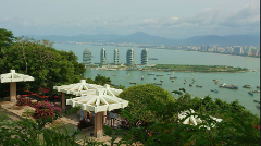A scenic view of Sanya, China, showcasing distinctive architectural buildings along the coastline from an elevated perspective with lush vegetation and white canopies in the foreground.