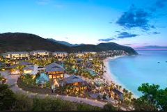 Here's a description of the image:A scenic coastal resort nestled between lush green hills and a pristine white sandy beach, with modern buildings illuminated against a twilight sky.