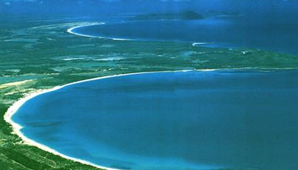 An aerial view showcases the coastline, with turquoise waters meeting sandy beaches and lush green landscapes under a blue sky.