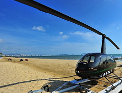 A black helicopter sits on a sandy beach under a blue sky, with a city skyline visible in the background.