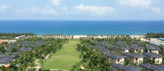 An aerial view showcases a tropical resort with buildings nestled among lush greenery, a pristine beach, and the vast blue ocean under a partly cloudy sky.