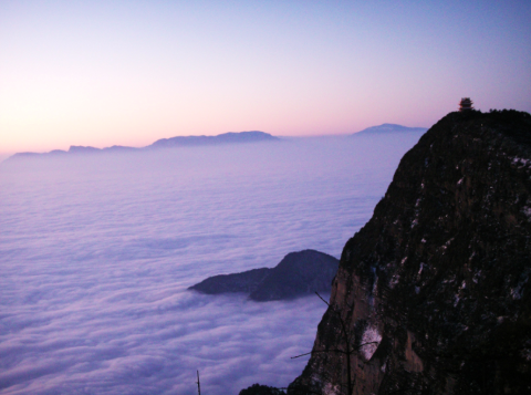 A scenic view from a mountain peak overlooks a sea of clouds at dawn, with distant mountain ranges and a small structure on the summit.