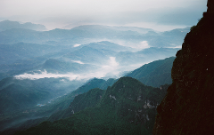 A scenic landscape of layered mountain ranges shrouded in mist, with a rocky cliff in the foreground.