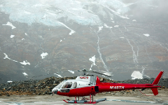 A red and white helicopter with the number N145TH sits on a rocky landscape with a glacier in the background under a cloudy sky.