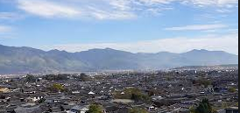 An aerial shot showcases a city with dark roofs nestled against a backdrop of hazy mountains under a light blue sky.