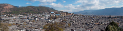 Panoramic view of a dense cityscape with gray-tiled roofs, nestled in a valley with mountains in the background under a partly cloudy sky.