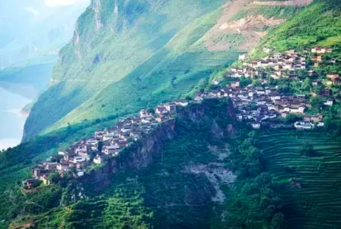 A village clings to a steep mountainside with terraced fields, offering a breathtaking vista of houses nestled in a verdant, hilly landscape beside a body of water.
