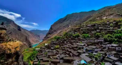 A village clings to the steep slopes of a mountain valley beneath a bright blue sky.