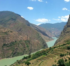 A scenic view of a wide river winding through a valley flanked by steep, terraced mountains under a blue sky with scattered clouds.