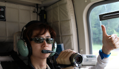 A woman with sunglasses and headset is filming with a JVC Everio HD camcorder inside a helicopter and giving a thumbs up.