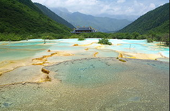 This is a vibrant, tiered landscape featuring turquoise and yellow mineral pools cascading down a slope in Huanglong, China, with lush green mountains and a traditional Chinese temple in the background.