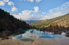 A serene mountain landscape features tiered turquoise pools reflecting the sky, nestled between forested slopes under a partly cloudy sky.