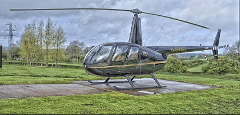 A black helicopter sits on a concrete pad surrounded by green grass, trees, and a cloudy sky.
