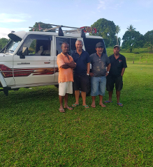 Four men are standing in front of a white Land Cruiser on a grassy field, with trees in the background.