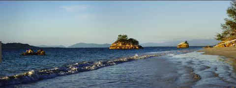A scenic view of a beach with small, rocky islands in the sea, framed by a clear sky and distant mountains.
