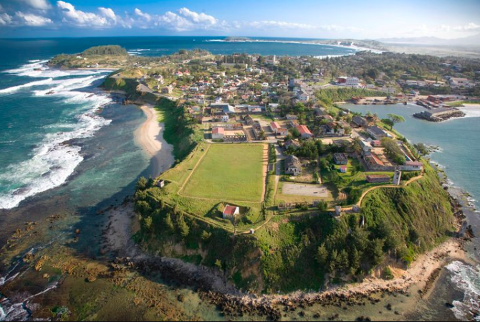 An aerial view captures the coastal town of Fort Dauphin, Madagascar, with a prominent fort on a cliffside, surrounded by vibrant blue ocean and lush green landscapes.