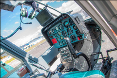 Cali Helicopter Tour - Best Value Aerial Tour - Best Pilots A cockpit view of a helicopter with a blue control panel, various gauges, a steering wheel, and a hanging headset, set against a backdrop of a partly cloudy sky and ground.