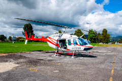 Cali Helicopter Tour - Best Value Aerial Tour - Best Pilots A white and red Bell 407 helicopter with the registration HK-3039 is parked on a gray asphalt landing pad with green grass and trees in the background, under a cloudy sky.
