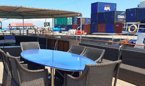 Outdoor seating with a blue table and wicker chairs on a deck, with a backdrop of colorful shipping containers and a bustling port scene under a sunny sky.