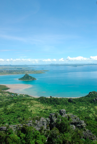 A scenic landscape view with lush green mountains in the foreground, a turquoise body of water with an island in the middle, and a clear blue sky with fluffy white clouds above.