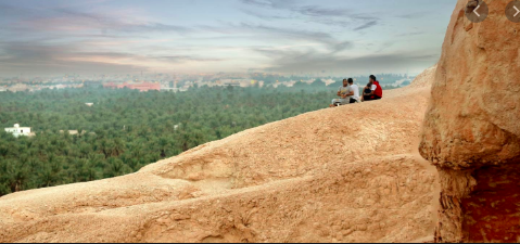 Three people sit on a rocky outcrop overlooking a lush green forest and distant cityscape under a cloudy sky.