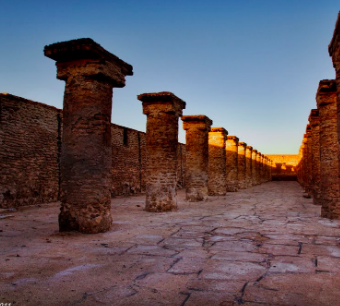 Here's a description of the image:A long row of weathered stone columns with square capitals stands in an ancient courtyard, bathed in the warm light of the setting sun, creating long shadows on the stone-paved ground.