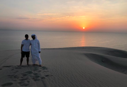 Two men stand on a sand dune overlooking the ocean during a sunset.