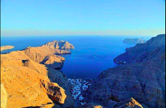 An aerial view showcases a bay surrounded by mountains, with a small village nestled between the peaks and clear blue skies above.