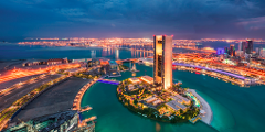 This stunning aerial shot captures a modern cityscape at twilight, featuring a tall skyscraper on an island surrounded by water, with bridges connecting it to the mainland and the distant city lights twinkling in the background.