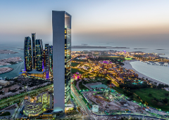 This is an aerial view of Abu Dhabi, with skyscrapers, illuminated buildings, a beach, and islands, all under a dusky sky.