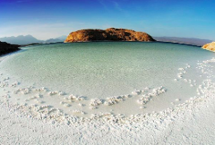 A wide-angle shot showcases a pristine white salt lake with an island in the background under a clear blue sky in Djibouti.