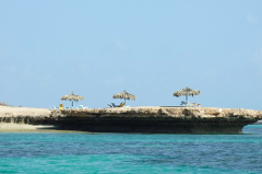 A picturesque view of a small, rocky island with three thatched umbrellas and beach chairs, surrounded by clear turquoise water under a clear blue sky.