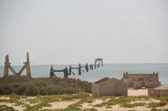 Com certeza! Aqui está uma descrição da imagem, dentro do limite de caracteres: A imagem retrata um píer em ruínas que se estende mar adentro a partir de uma praia arenosa com vegetação rasteira, exibindo vestígios de edifícios e estruturas antigas.