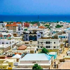 Aqui está uma descrição da imagem:Uma vista panorâmica de uma cidade na Somália, provavelmente perto da costa, mostrando edifícios próximos uns dos outros com uma mistura de telhados planos e inclinados, com o oceano visível à distância.