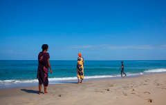 Três pessoas em uma praia de areia: uma mulher em pé na beira da água com um vestido amarelo, outra mulher em pé mais adiante na praia com um vestido escuro e um menino caminhando pela orla. O mar está azul e o céu está limpo.