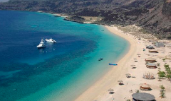 An aerial view of a picturesque beach with turquoise waters, boats, huts, umbrellas, and mountainous terrain in the background.
