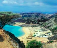 A scenic view of a turquoise bay nestled between arid hills and a sandy beach, with a white building complex nearby, all under a cloudy sky in Djibouti.