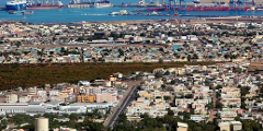 This is a high-angle view of Djibouti City, showing a sprawling urban landscape near the sea, with ships and port facilities visible in the distance.
