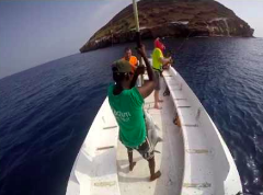 En vidvinkelbild föreställer tre personer som fiskar från en båt i Djibouti, där en person håller upp en fisk de fångat, mot en liten, klippig ö i bakgrunden.