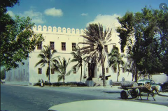 A vintage view shows a white, fortress-like building with battlements, palm trees, and a horse-drawn cart on the street in front, likely in Mogadishu, Somalia.