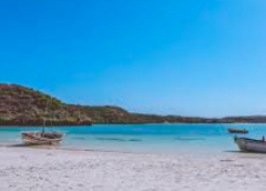 Este cenário costeiro sereno apresenta uma praia de areia branca imaculada com barcos ancorados, águas azul-turquesa e um cenário de terreno montanhoso sob um céu azul claro.