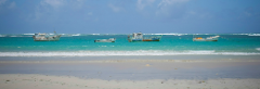 Uma cena serena de praia apresenta águas azul-turquesa, ondas suaves e quatro pequenos barcos parados perto da costa sob um céu parcialmente nublado.
